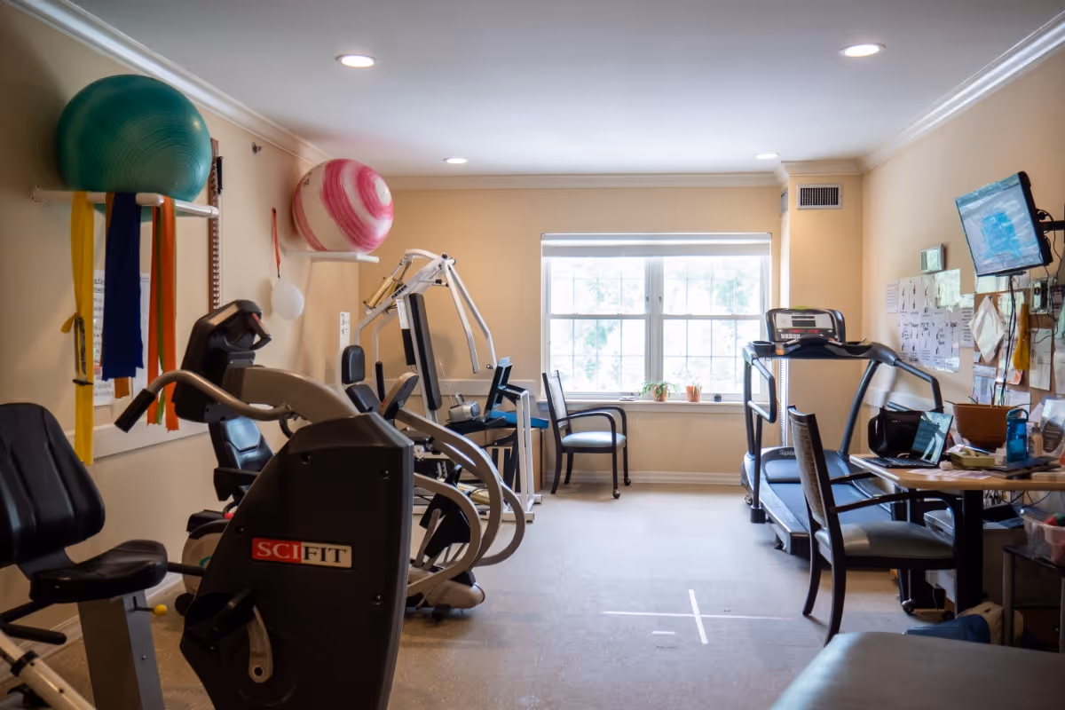 A small fitness room with exercise equipment including a recumbent bike, a multi-exercise machine, and a treadmill. Two large exercise balls are mounted on the wall with colorful resistance bands hanging from the rack. There is a window letting in natural light, a couple of chairs near the window, and a table with a laptop and other items on the right side of the room.