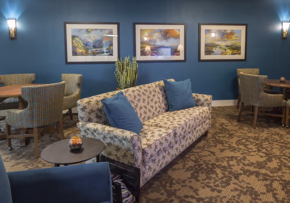 Comfortable common room featuring a floral-patterned sofa with blue pillows, round tables and upholstered chairs against a blue wall with three framed landscape prints.