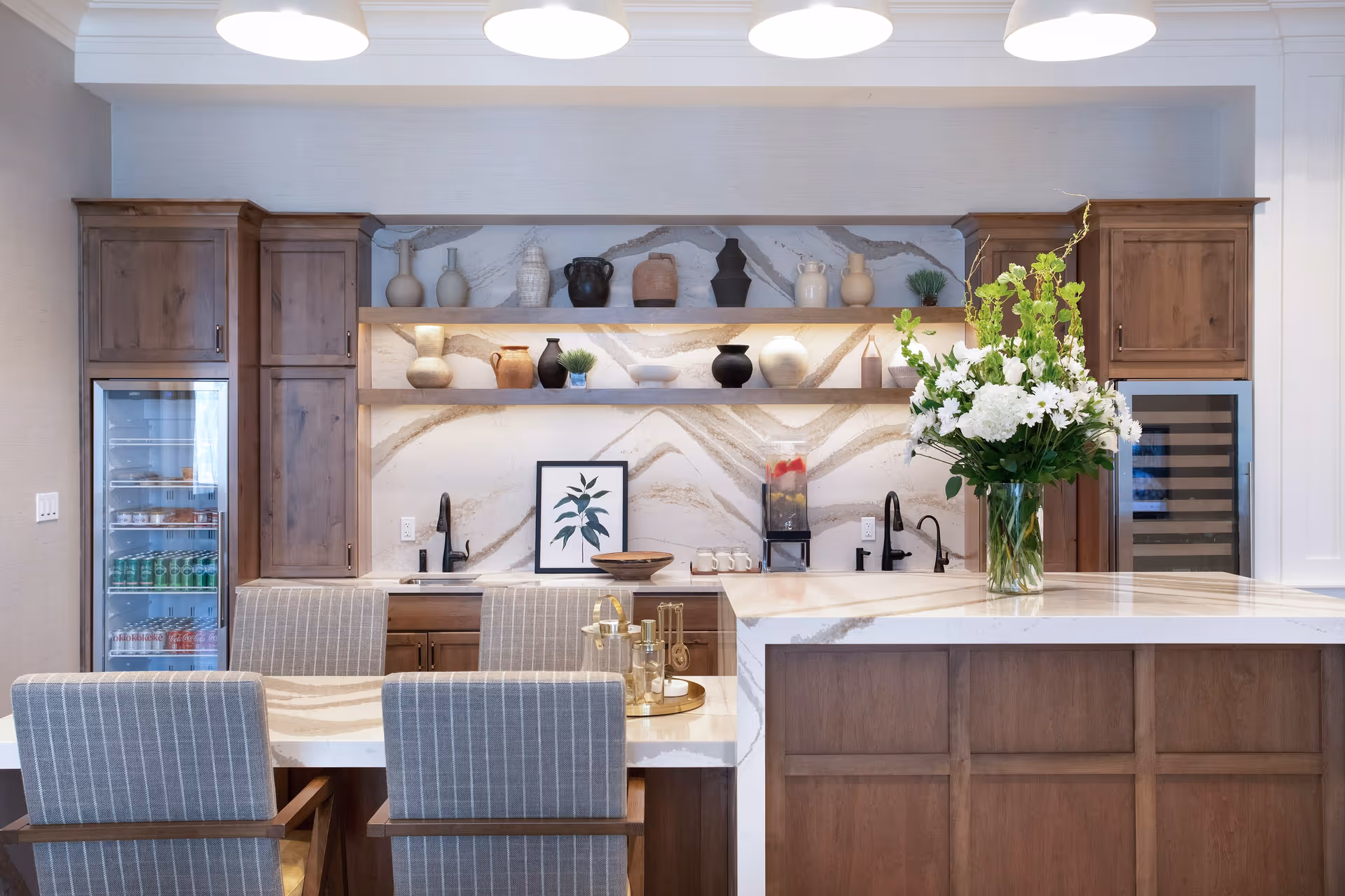 A modern kitchen area with wooden cabinets and marble countertops. There are two built-in refrigerators on either side, open shelves with decorative pottery and plants, and a large vase with white flowers on the island. Four upholstered chairs with striped fabric are positioned at the counter. The space is well-lit with three overhead pendant lights.