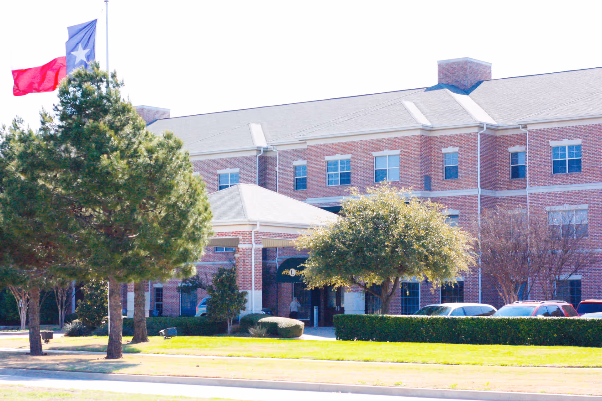 Exterior view of a large brick building with multiple windows, a covered entrance, and a parking area with several cars. There are trees and well-maintained bushes in front of the building, and a Texas state flag is flying on a flagpole to the left.