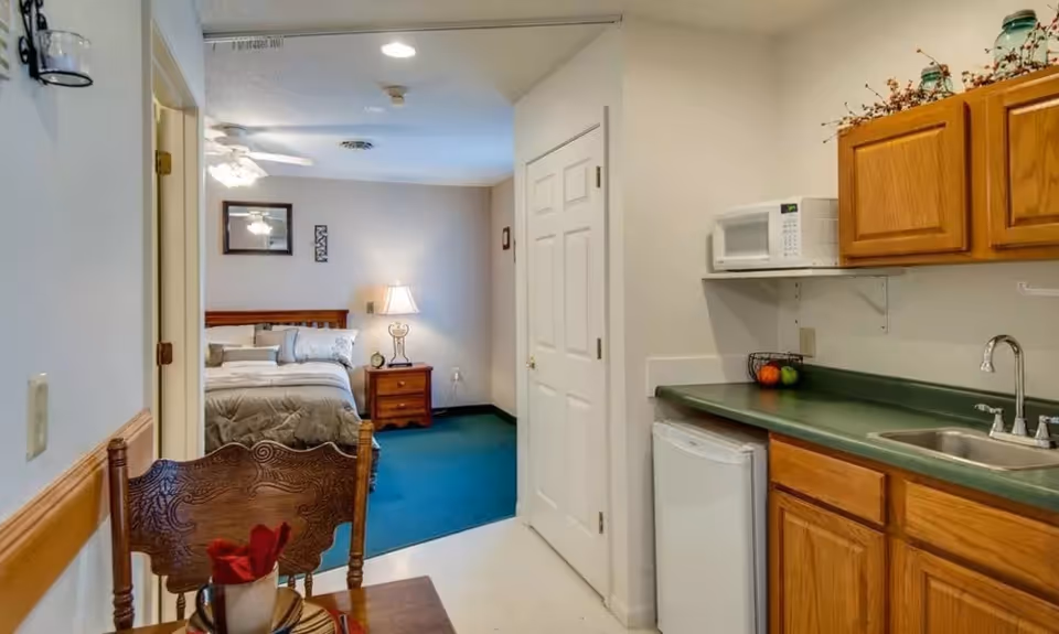 View of a small living space in a senior living facility showing a kitchenette with wooden cabinets, a sink, a microwave, and a mini refrigerator. Adjacent to the kitchenette is a dining table with a wooden chair and a red napkin. In the background, there is a bedroom with a bed, nightstand, lamp, and wall decorations.