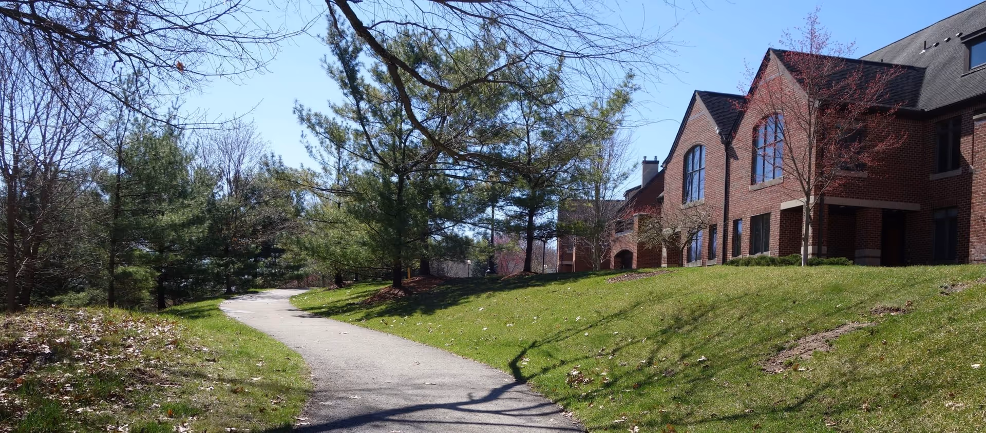 A paved walking path curves through a grassy area with trees on both sides. To the right, there is a large brick building with multiple windows and a sloped roof. The sky is clear and blue, indicating a sunny day.