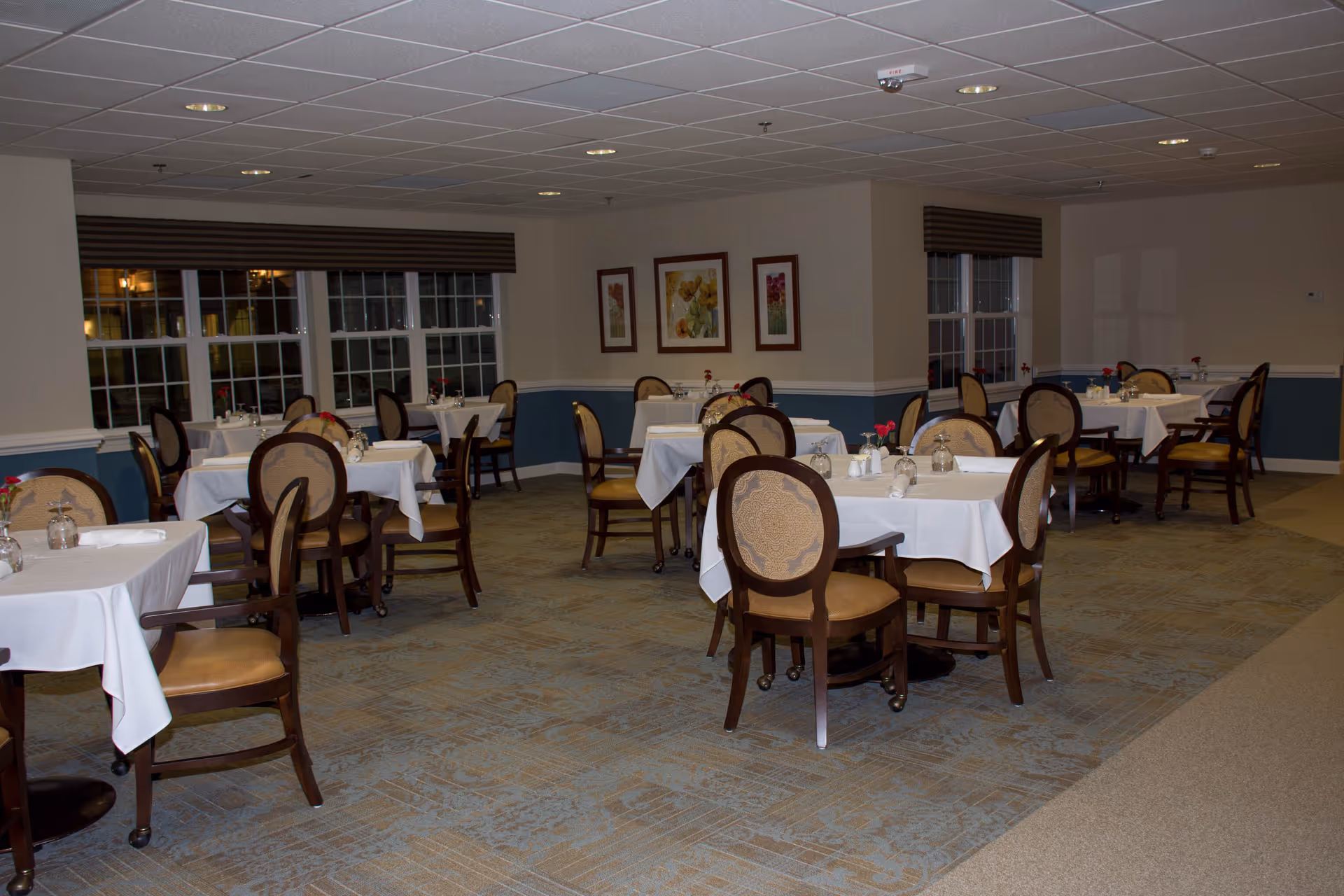 A dining room with multiple tables covered in white tablecloths, each set with glasses, napkins, and small flower vases. The room has large windows with brown valances, framed floral artwork on the walls, and carpeted flooring.