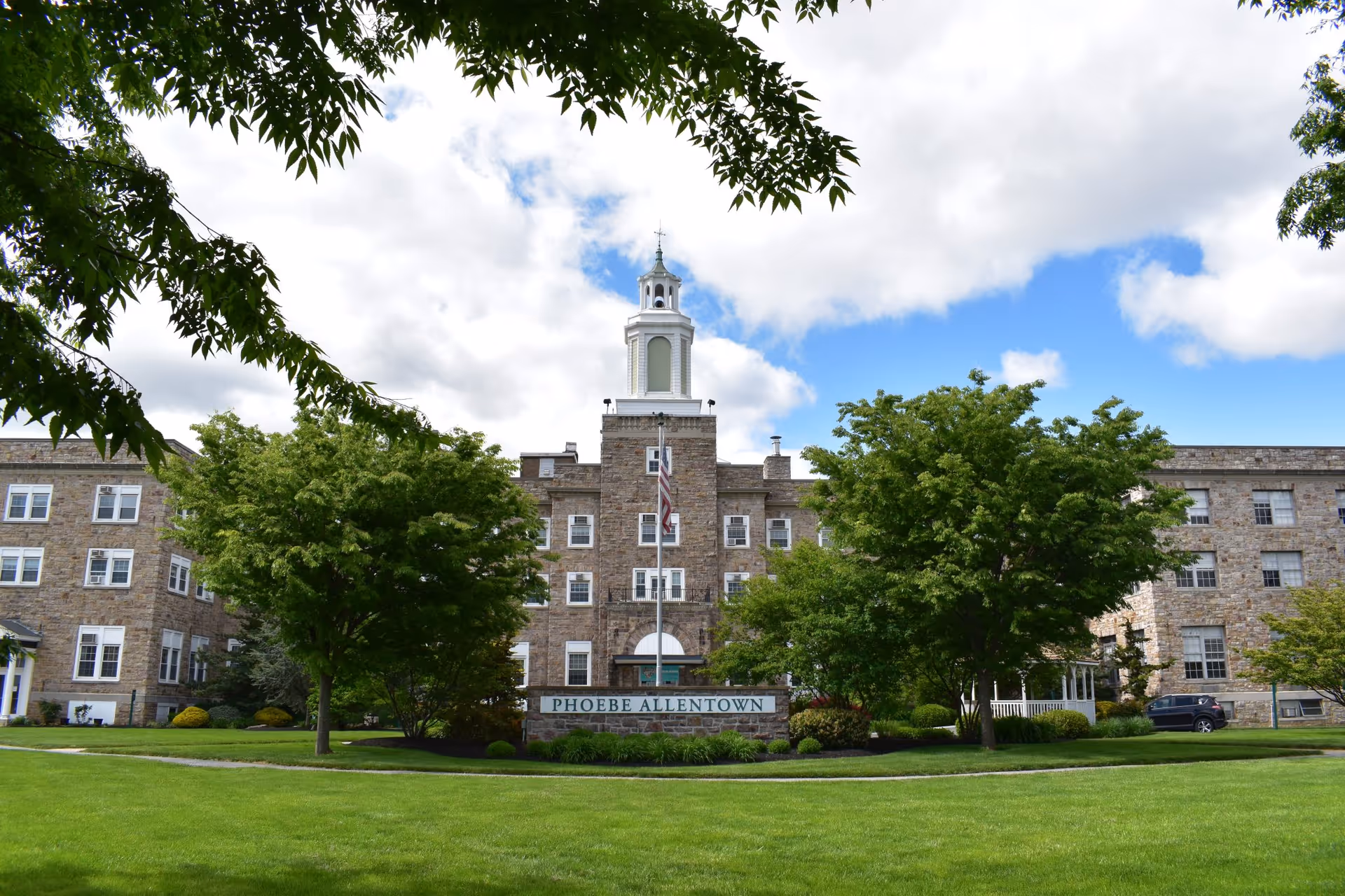Front exterior view of a large stone building with multiple windows, a central tower with a cupola, and a flagpole in front. The building is surrounded by green trees and a well-maintained lawn under a partly cloudy sky. A sign in front reads 'PHOEBE ALLENTOWN'.