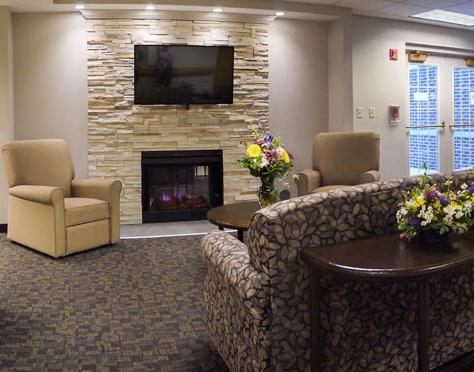 A comfortable common lounge with armchairs and a patterned sofa facing a stone-clad fireplace topped by a wall-mounted TV, with floral arrangements on tables.