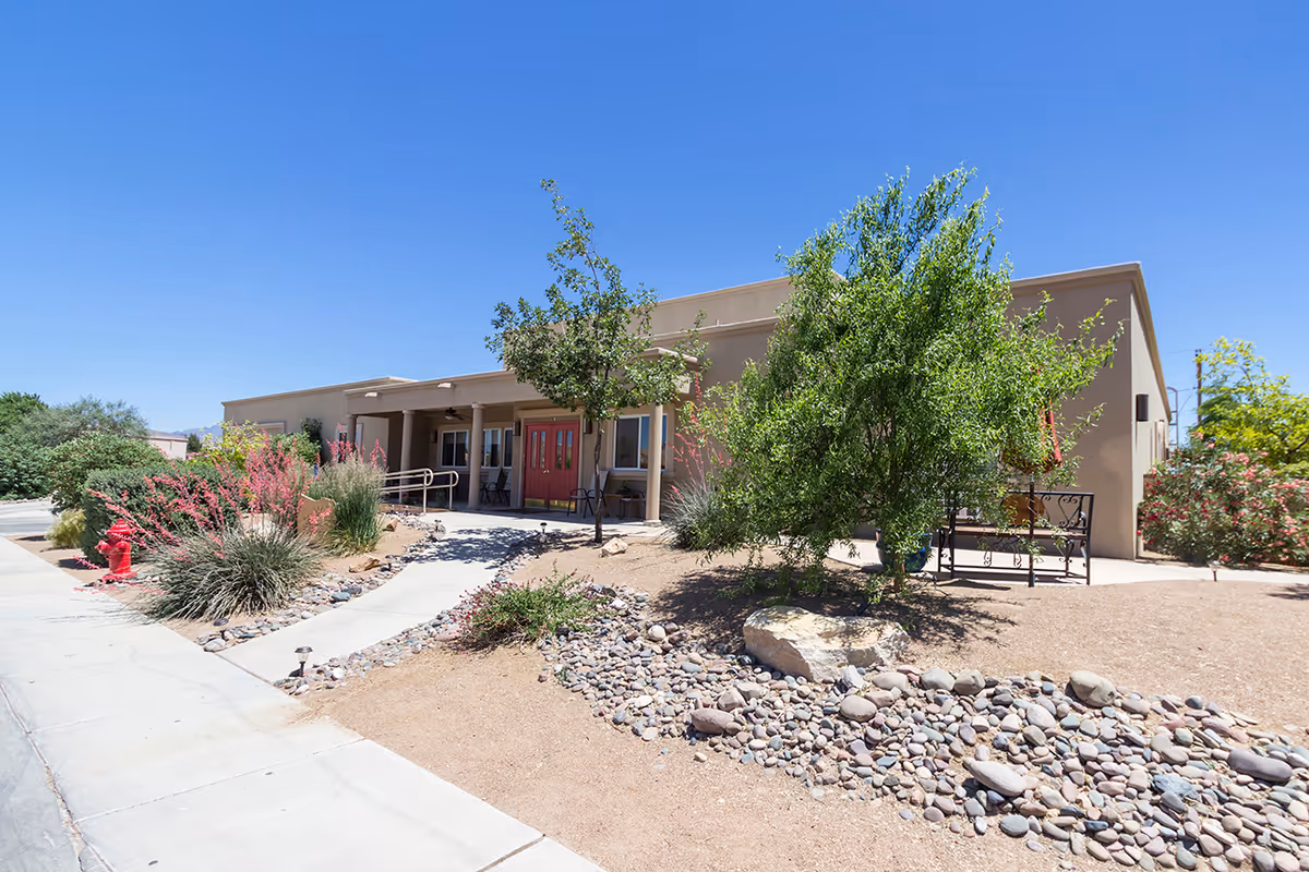 Exterior view of a single-story building with a beige facade under a clear blue sky. The entrance features double red doors with a covered porch and a wheelchair ramp. The surrounding landscape includes desert-style plants, small trees, rocks, and a concrete walkway leading to the entrance.