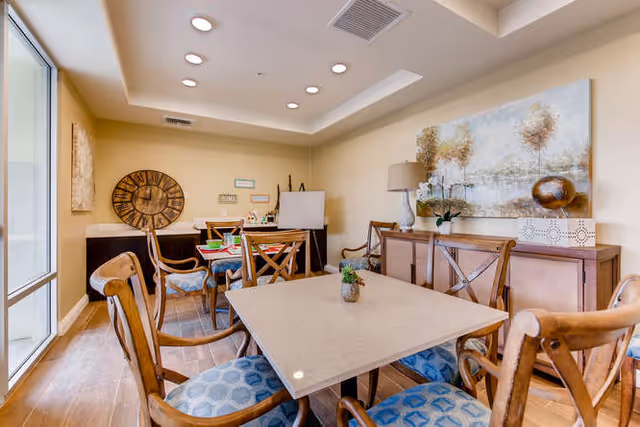 A well-lit interior room with a square table surrounded by wooden chairs with blue patterned cushions. The room features a large wall clock, a sideboard with a lamp and decorative items, and a large painting of trees on the wall. There is a glass door on the left side allowing natural light to enter.