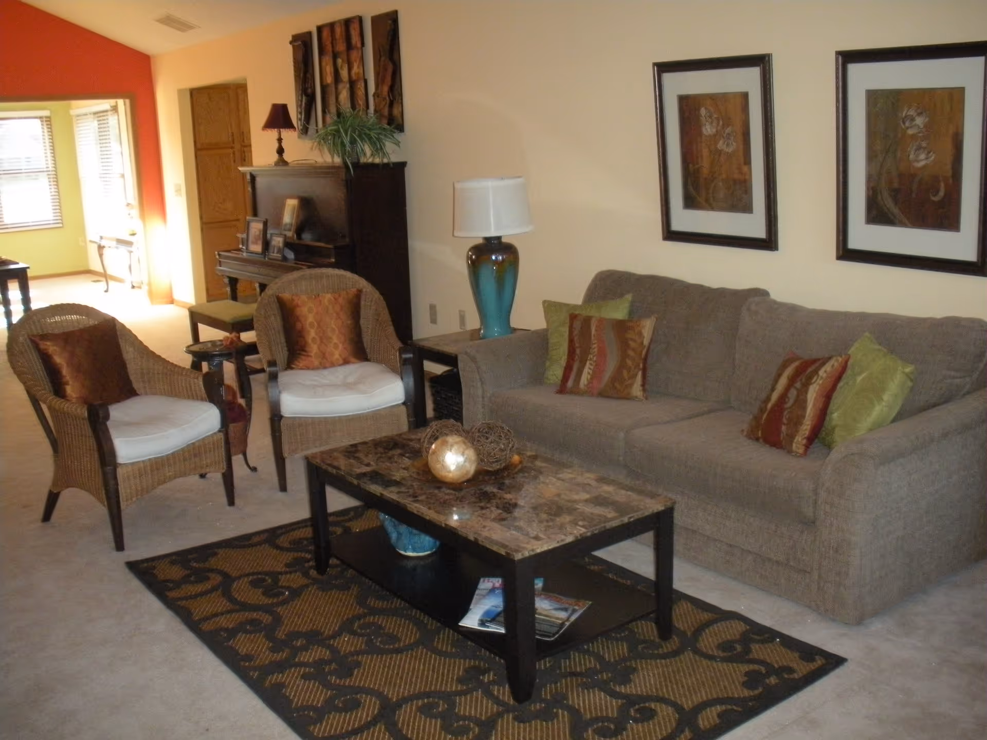A cozy living room with a gray sofa adorned with multiple decorative pillows, two wicker armchairs with cushions, a marble-top coffee table with decorative items, a patterned rug underneath, a lamp on a side table, framed floral artwork on the wall, and a piano with a lamp and plant on top in the background.