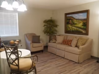 A cozy living room area with a beige sofa adorned with multiple cushions, a cushioned armchair, a round table with two chairs, a potted plant in the corner, and a large framed landscape painting on the wall. The room has wood flooring and a window with blinds.