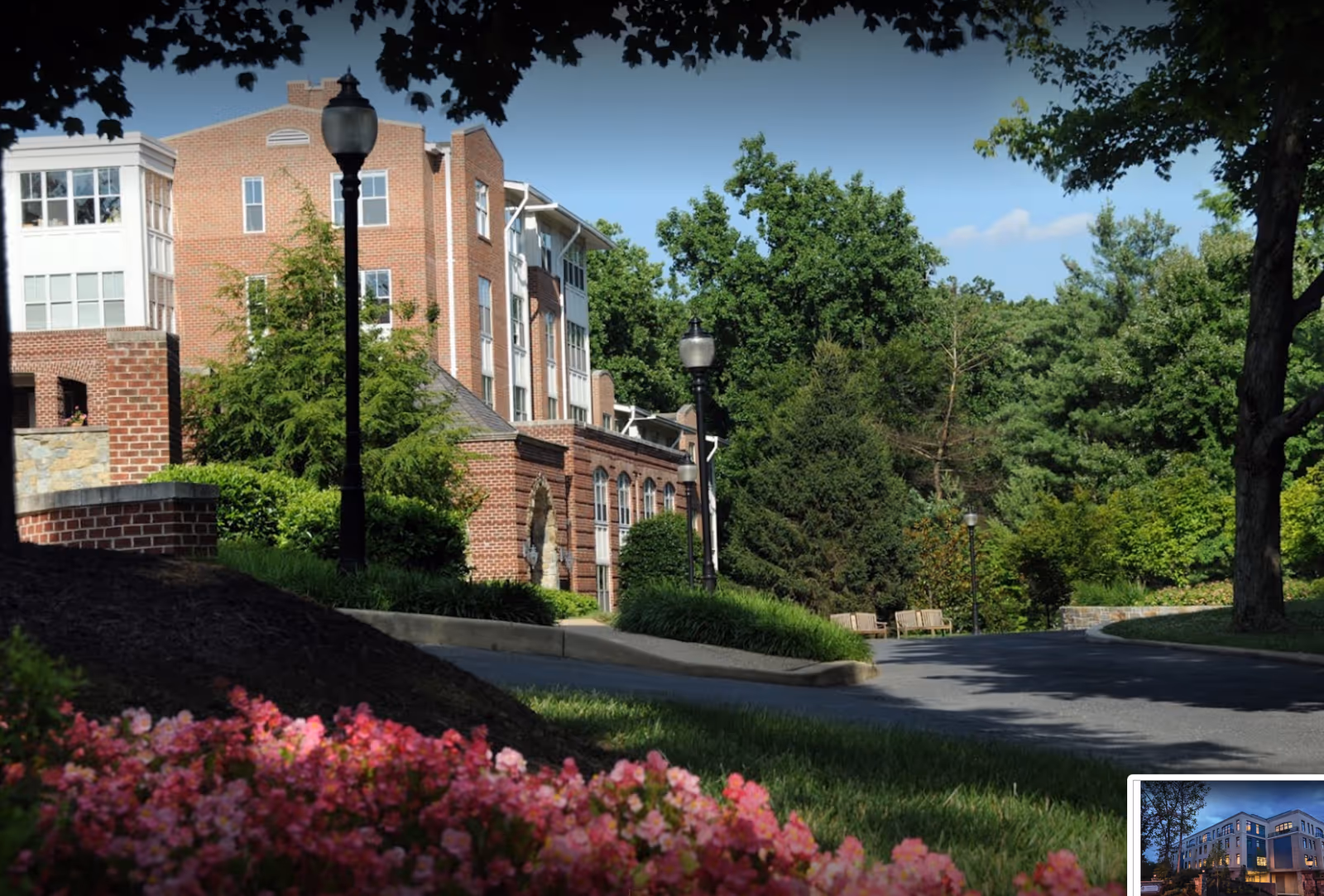 Brick senior living building with lamp posts, trees, a curved driveway, and flowering shrubs in the foreground.