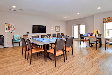 A spacious, well-lit common area with multiple tables and chairs arranged for group activities. The room has light wood flooring, neutral-colored walls, a wall-mounted flat-screen TV, and framed artwork. There are large windows and glass doors letting in natural light, and some tables have craft supplies and flowers on them.