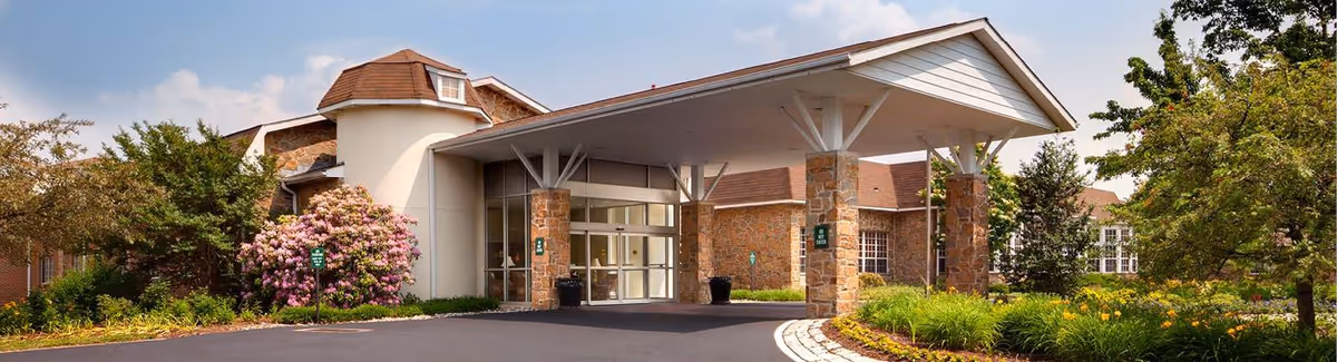 Front entrance of Normandy Farms Estates with a covered porte-cochere supported by stone columns, landscaped beds, and the building facade.