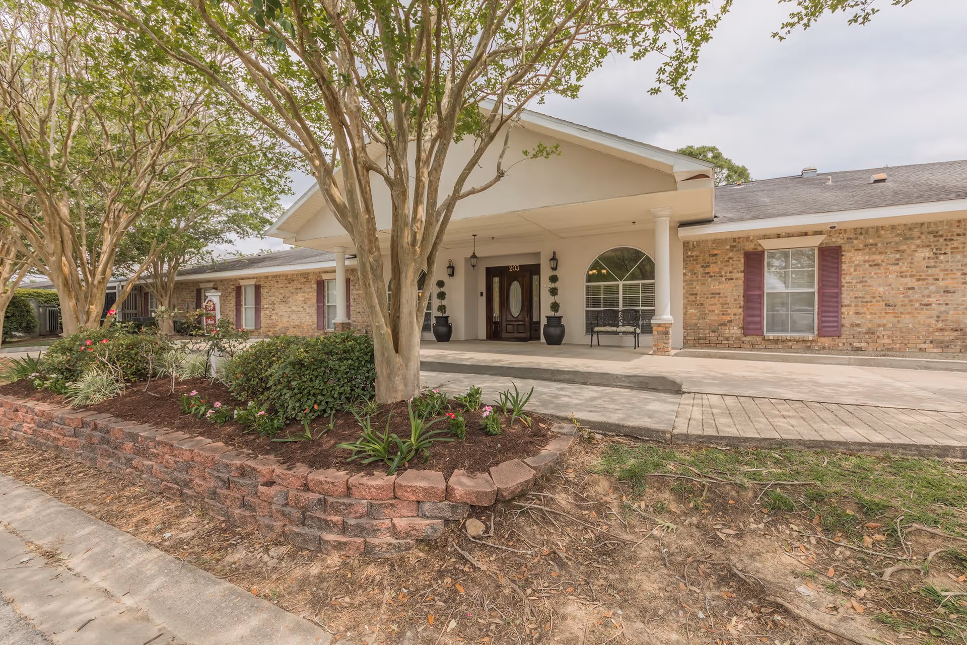 Front exterior view of a single-story brick building with a covered entrance supported by white columns. There are two large potted plants flanking the dark wooden front door with an oval glass panel. The building has several windows with purple shutters and a small bench near one window. In front of the building is a landscaped area with trees, bushes, and flowers bordered by a low brick retaining wall.
