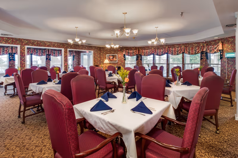 Dining room with multiple tables set with white tablecloths, navy napkins, floral centerpieces, and burgundy upholstered chairs.