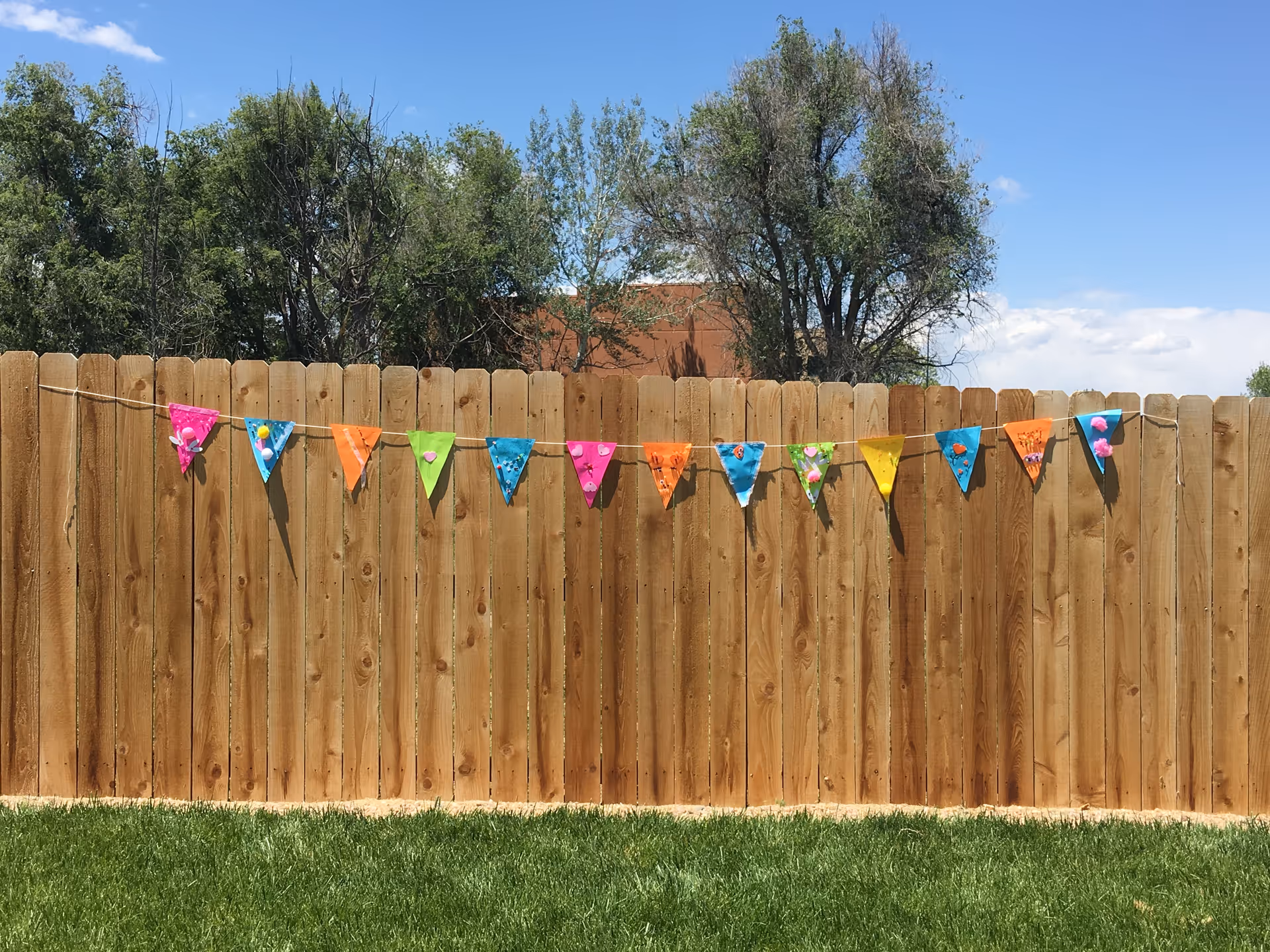 A wooden backyard fence decorated with colorful triangular pennant bunting, with grass in the foreground and trees and blue sky behind.