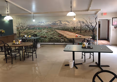 Dining room with several tables and chairs and a large mural of a country landscape on the back wall.