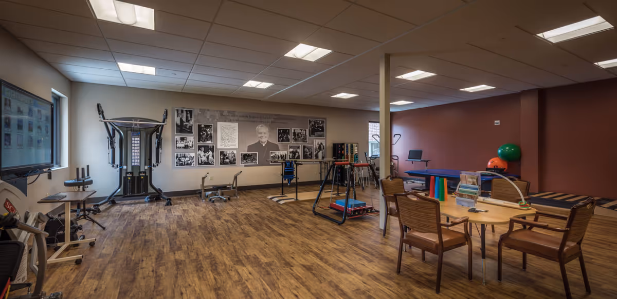 A spacious rehabilitation or therapy room with wooden flooring, exercise equipment, therapy tables, chairs around a table with therapy tools, and a wall decorated with black and white photos and a large portrait. The room is well-lit with ceiling lights and has a window on the left side.