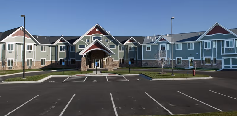 Front exterior of a two-story green and stone senior living building with a covered entrance and an empty parking lot under a clear sky.