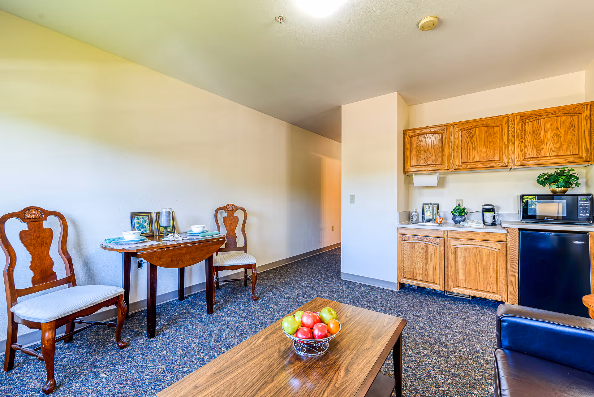 Bright studio-style living area with a small kitchenette, wooden cabinets, a two-seat dining table, and a coffee table with a bowl of fruit.