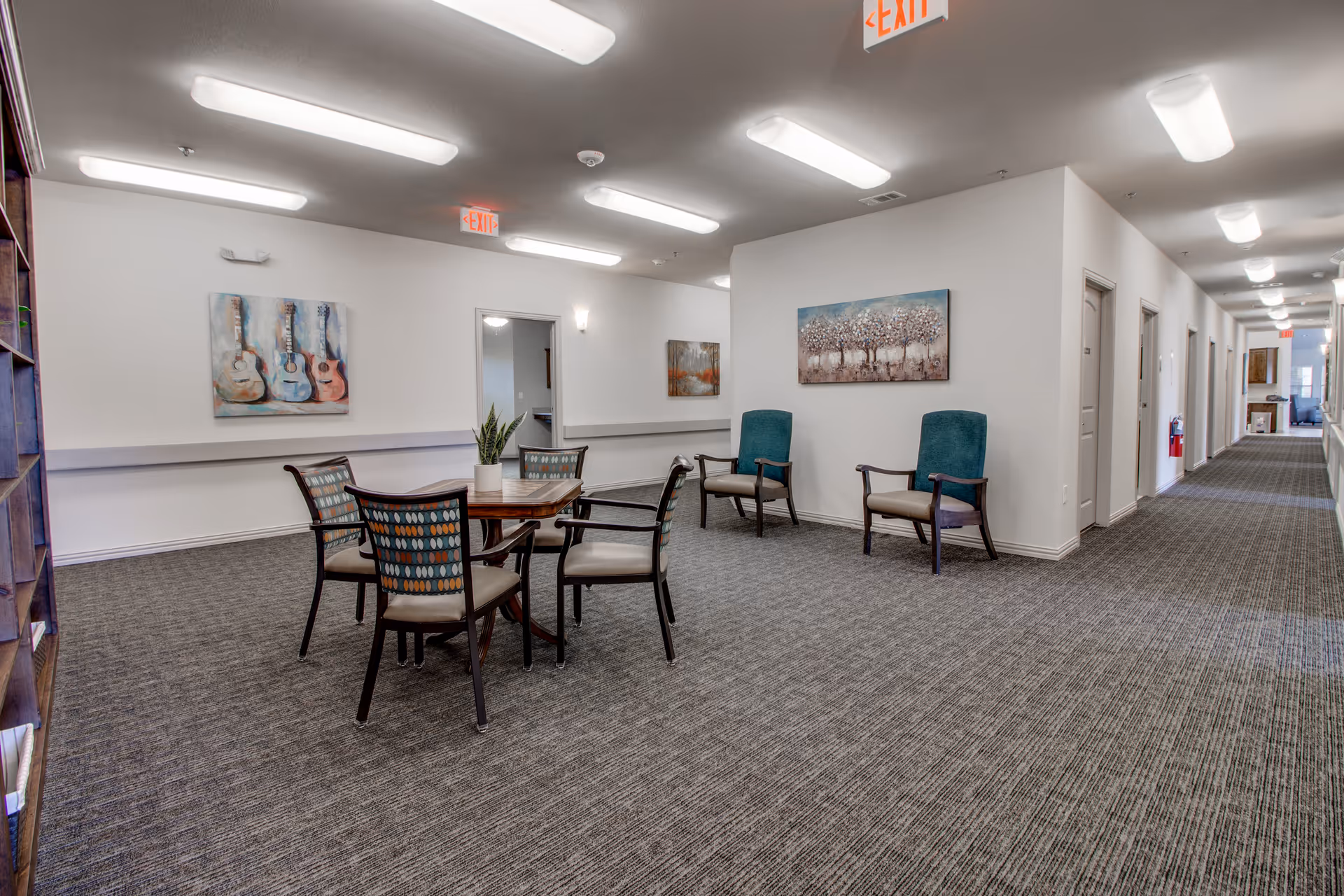 A spacious interior common area in an assisted living facility featuring a small wooden table with four chairs, two additional armchairs against the wall, carpeted floor, white walls adorned with paintings, and bright overhead lighting. A long hallway with multiple doors extends to the right.