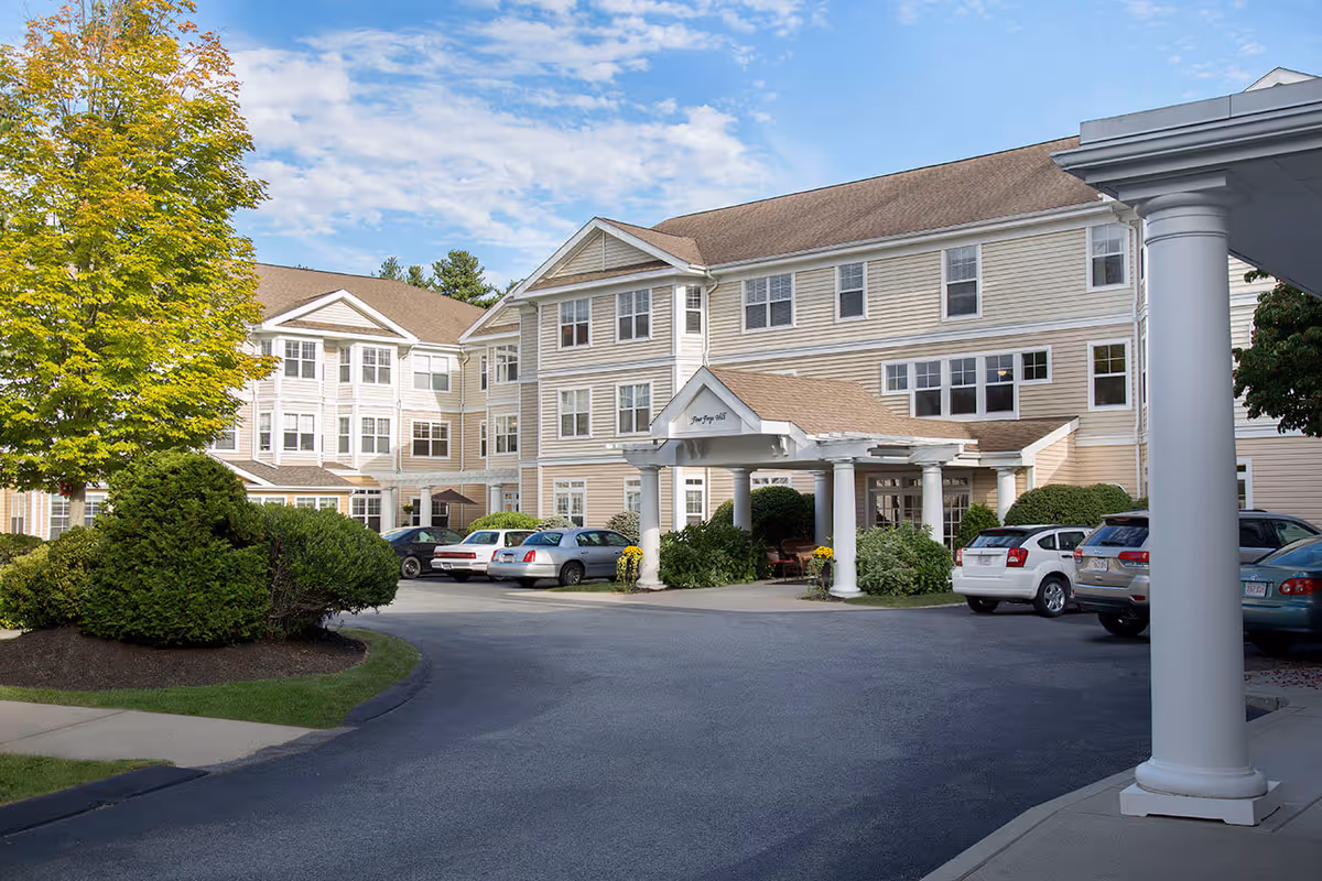 Exterior view of Benchmark Senior Living at Forge Hill, showing a large beige multi-story building with white trim and a covered entrance supported by white columns. Several cars are parked in front of the building, and there are green bushes and a tree with yellow-green leaves near the driveway under a partly cloudy blue sky.