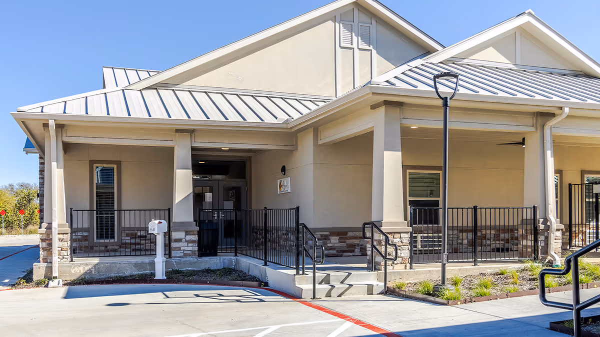 Front entrance of a beige senior living building with a covered porch, steps and ramp, black railings and a mailbox.