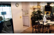 Interior view of a dining area in a retirement community with a glass-top dining table and chairs, a large plant, and a kitchen area visible in the background. There is a clock on the wall and a window with curtains letting in natural light.