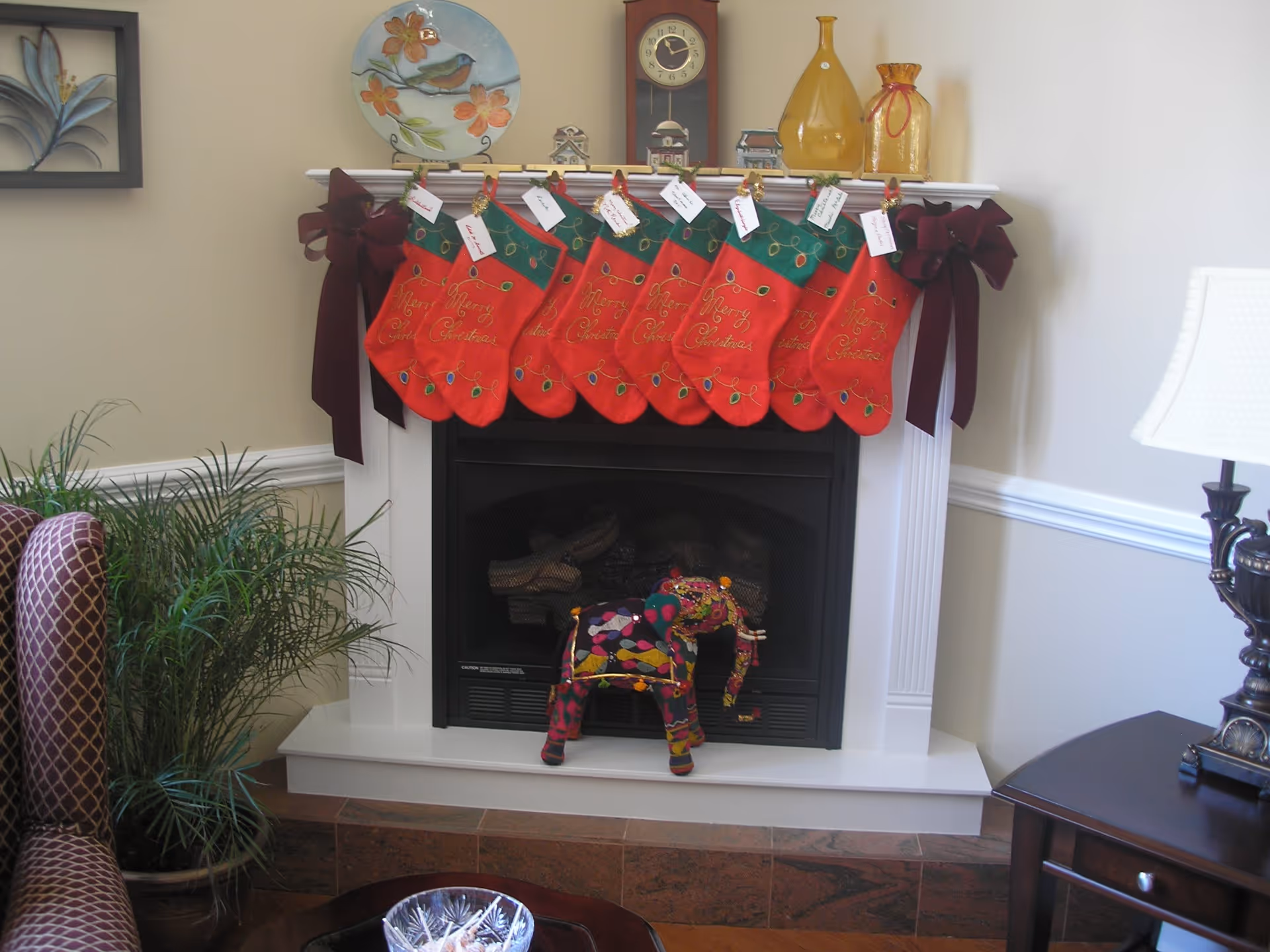 A cozy living room fireplace decorated for Christmas with seven red stockings hanging from the mantel, each with a name tag. The mantel also holds decorative items including a clock, a plate with a bird design, and two yellow glass bottles. A colorful stuffed elephant toy is placed in front of the fireplace. There is a patterned armchair and a green plant to the left, and a dark wooden side table with a lamp to the right.