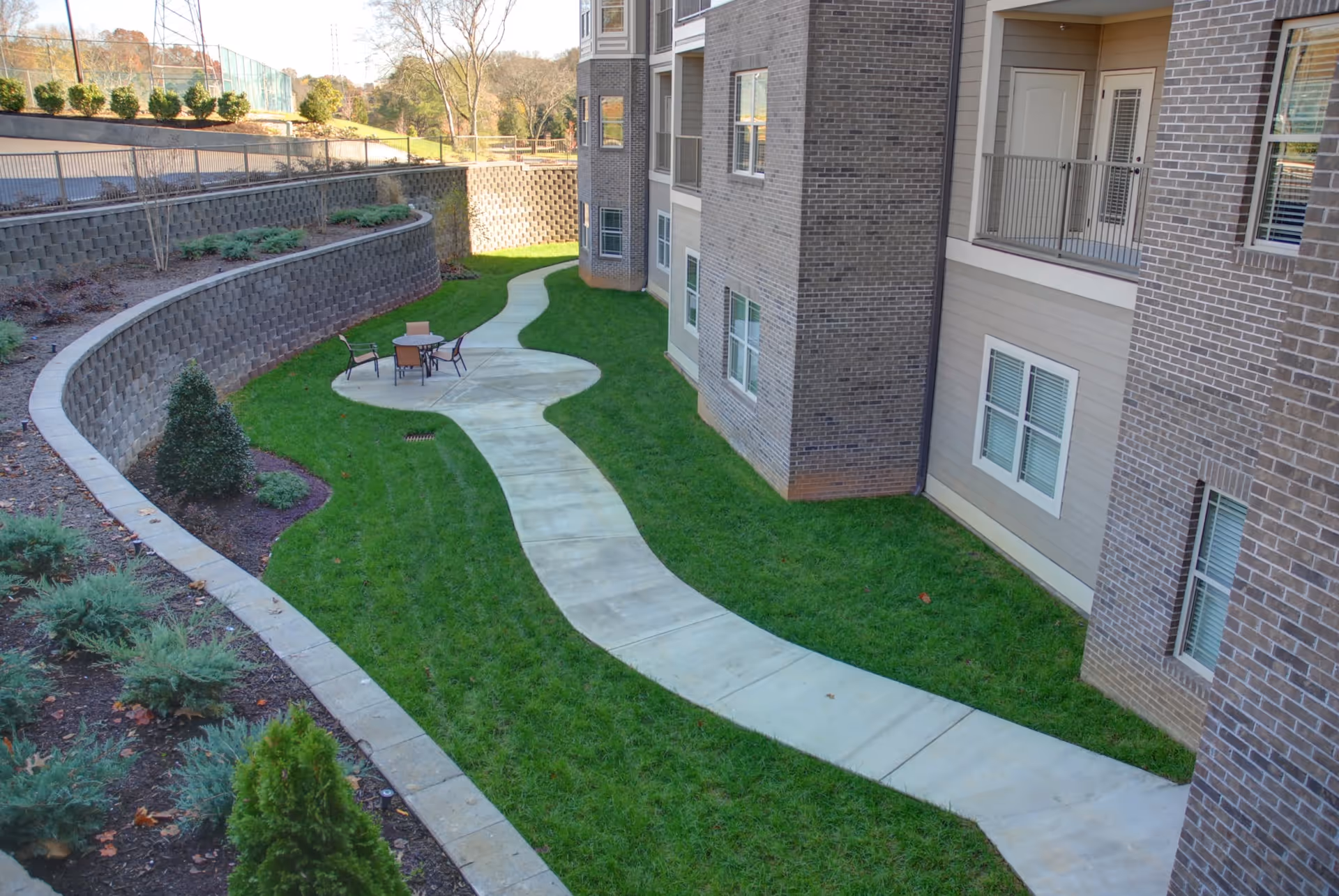 Outdoor view of a landscaped area at Rain Forest Landings with a curved concrete pathway leading to a small circular patio with a table and four chairs. The area is bordered by a retaining wall with shrubs and small trees, and the side of a multi-story brick building with windows and balconies is visible.