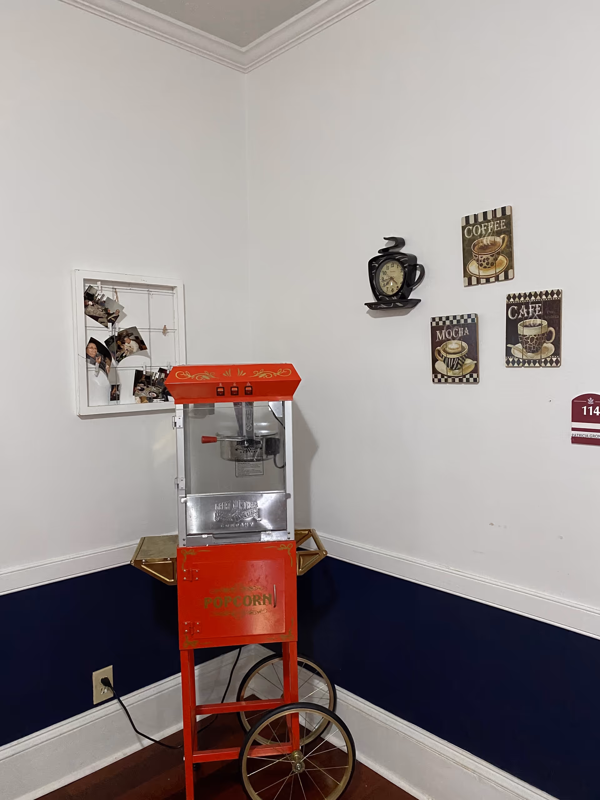 A corner of a room with a vintage-style red popcorn machine on wheels. The walls are white with a dark blue lower half. On the wall above the popcorn machine, there are three decorative coffee-themed plaques and a black clock shaped like a coffee cup. To the left, there is a white photo display frame with several photos clipped to it.
