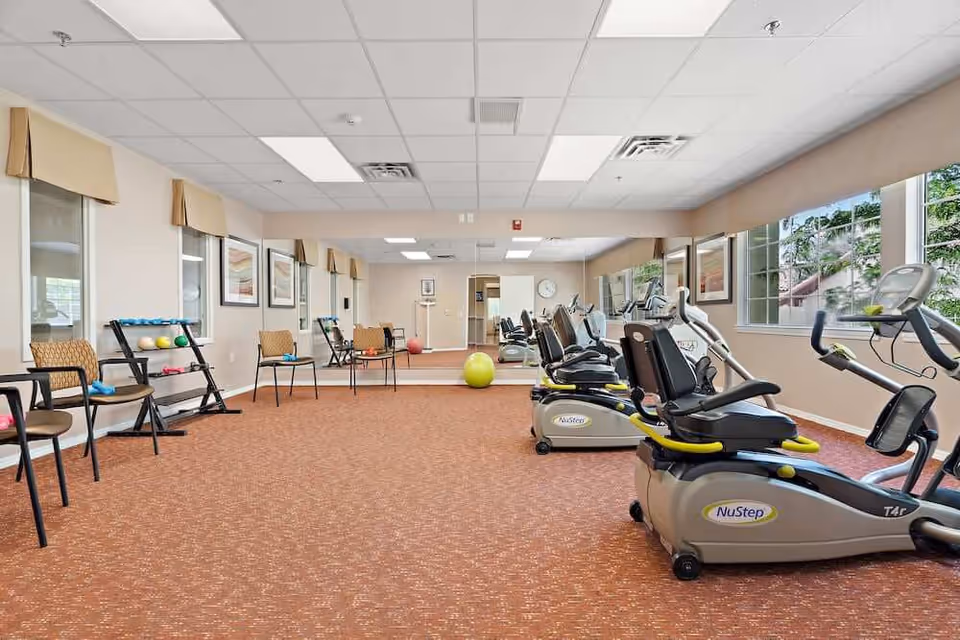 A fitness room with several NuStep exercise machines lined up on the right side, chairs and small exercise equipment like dumbbells and exercise balls on the left side, large mirrors on the far wall, and windows letting in natural light.