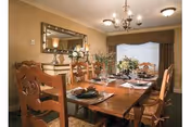 A warmly lit dining room with a wooden dining table set for six people. The table is decorated with floral centerpieces and place settings including plates, glasses, and napkins. The room features a large mirror on the wall, a chandelier overhead, and a window with curtains in the background.