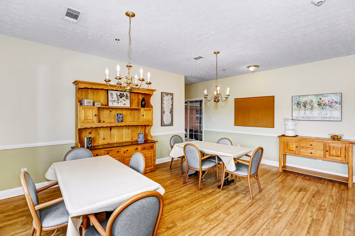 A dining room with two tables covered with white tablecloths, each surrounded by four chairs with wooden frames and gray cushions. The room has light green and white walls, wooden flooring, and two brass chandeliers hanging from the ceiling. There is a wooden hutch with decorative items and framed artwork on the walls, as well as a wooden sideboard with a glass pitcher and a bowl of fruit.