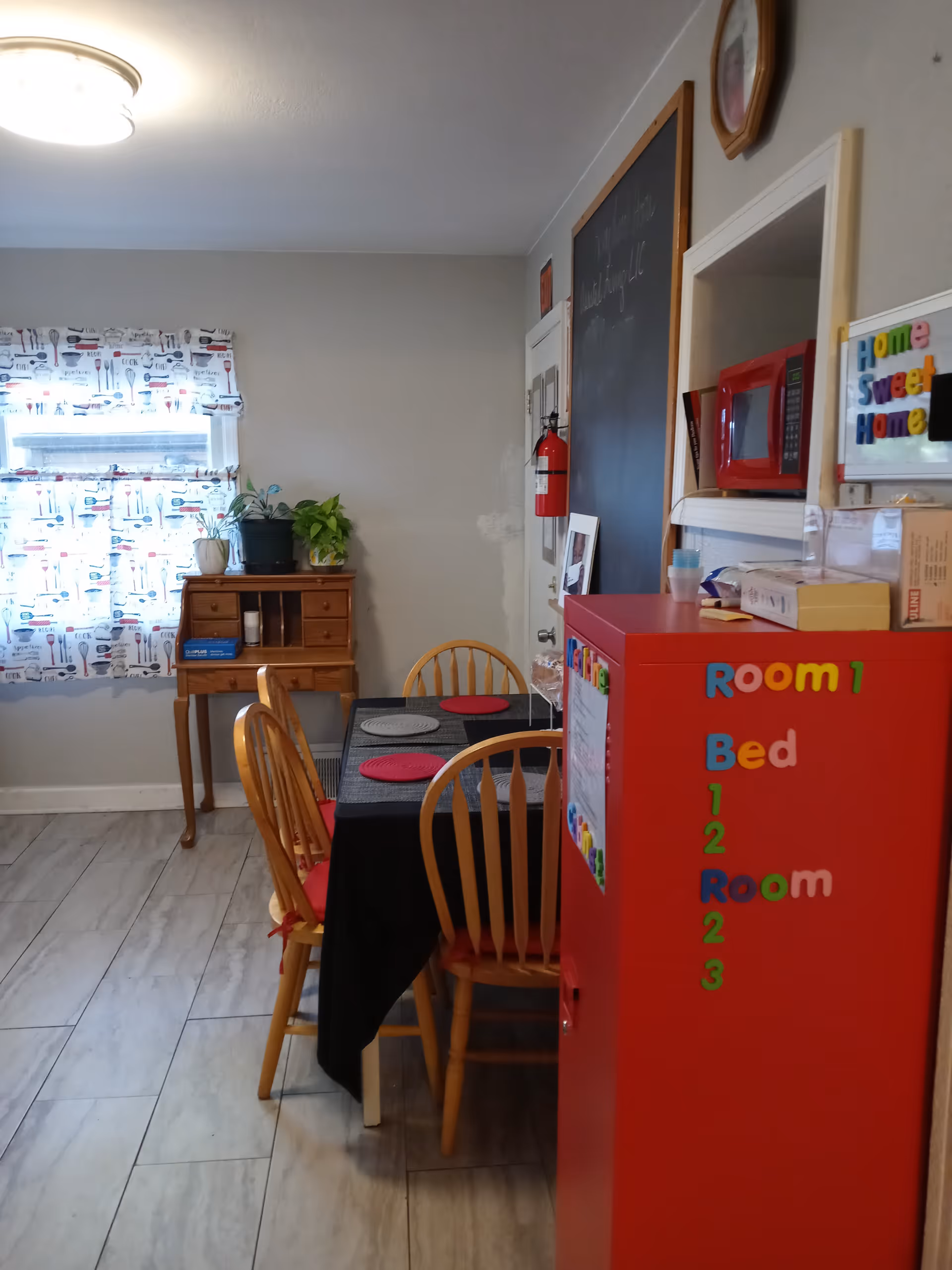 A small dining area with a table covered by a black tablecloth and four wooden chairs. The table is set with placemats. To the right, there is a red cabinet with colorful magnetic letters spelling out room and bed numbers. A small wooden desk with plants on top is positioned against the wall under a window with patterned curtains. A fire extinguisher is mounted on the wall near a door, and a chalkboard is visible on the wall above the cabinet. The floor is tiled with light-colored tiles.