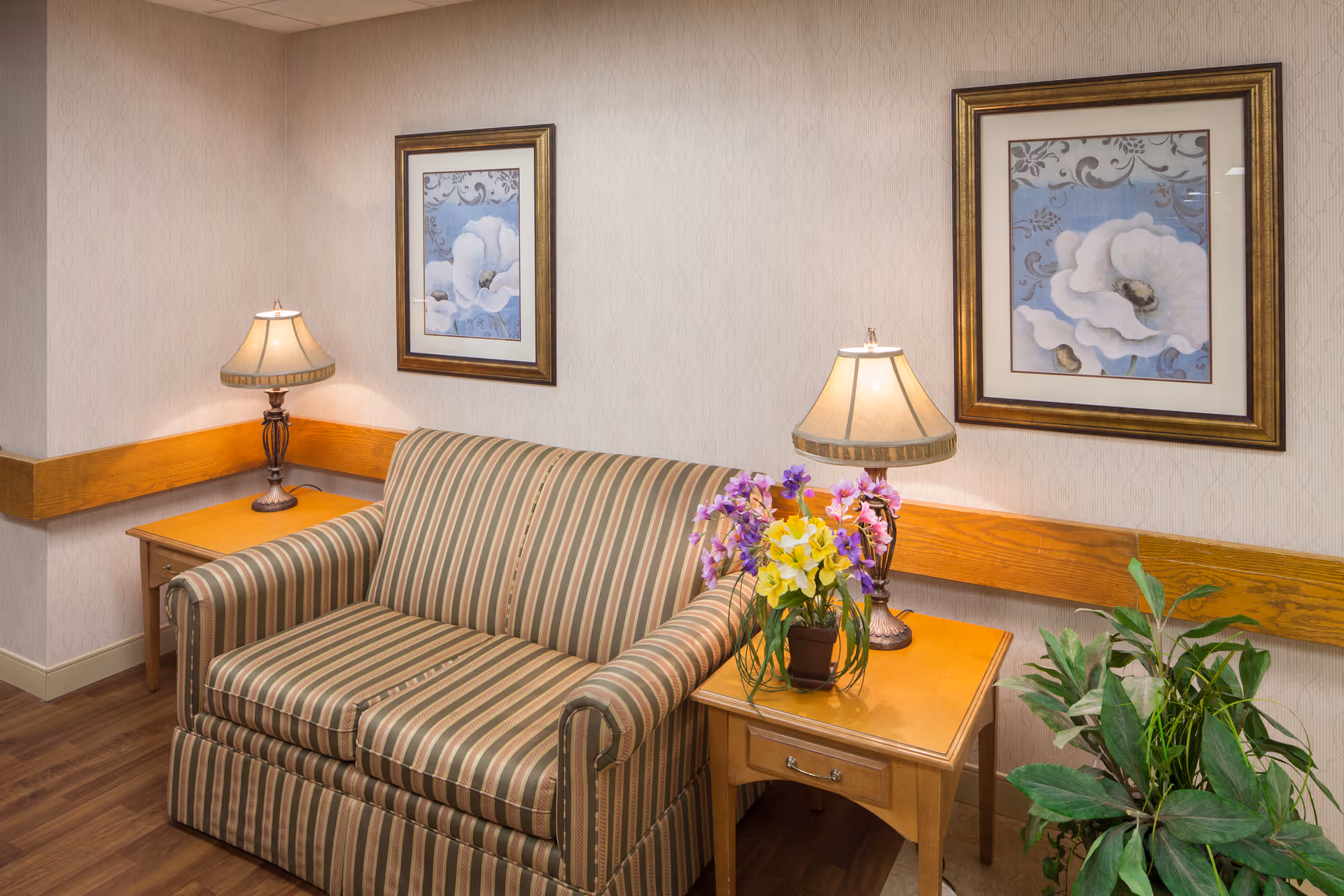 A striped loveseat between two wooden side tables with lamps, framed floral paintings on the wall, and potted flowers and plants in a lounge area.