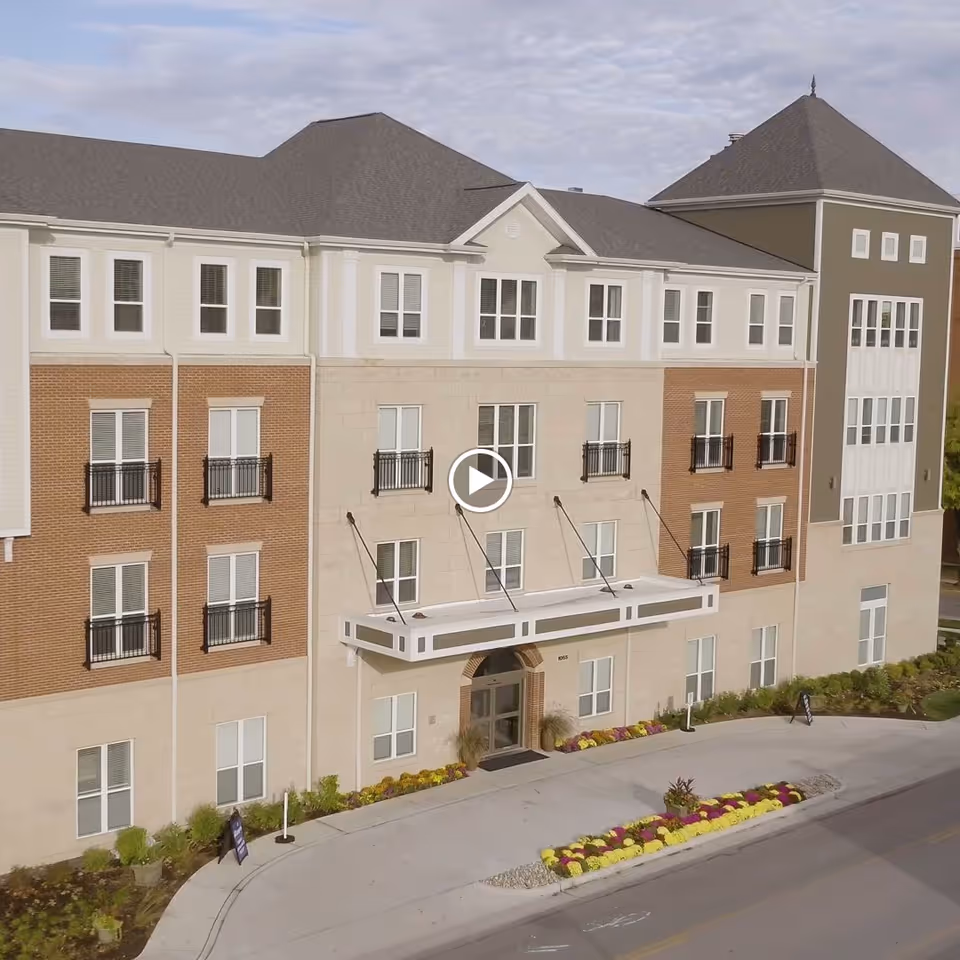 Exterior view of a multi-story senior living facility building with a combination of brick and light-colored siding. The entrance is covered by a white awning supported by metal rods. There are several windows with black railings and a landscaped area with flowers and shrubs along the sidewalk.