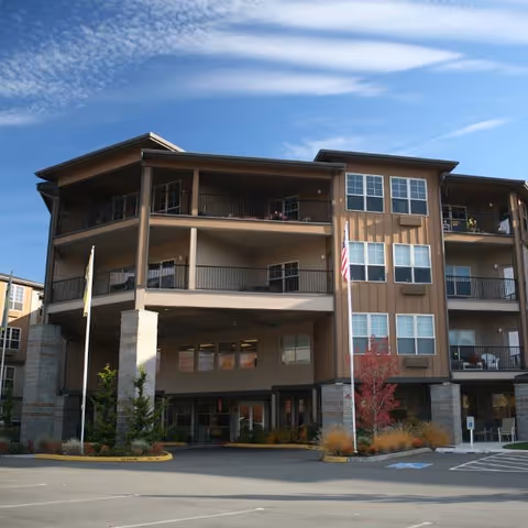Exterior view of a multi-story senior living facility building with balconies, large windows, and an entrance area. The building is surrounded by a parking lot, small trees, and landscaping under a partly cloudy sky.