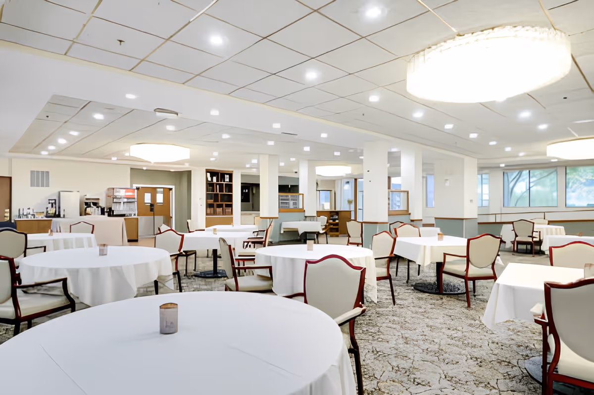 A spacious dining room with round and square tables covered in white tablecloths, surrounded by cushioned chairs with wooden frames. The room has a patterned carpet, large windows letting in natural light, and ceiling lights with large circular fixtures. There is a beverage station with coffee machines and other equipment in the background.