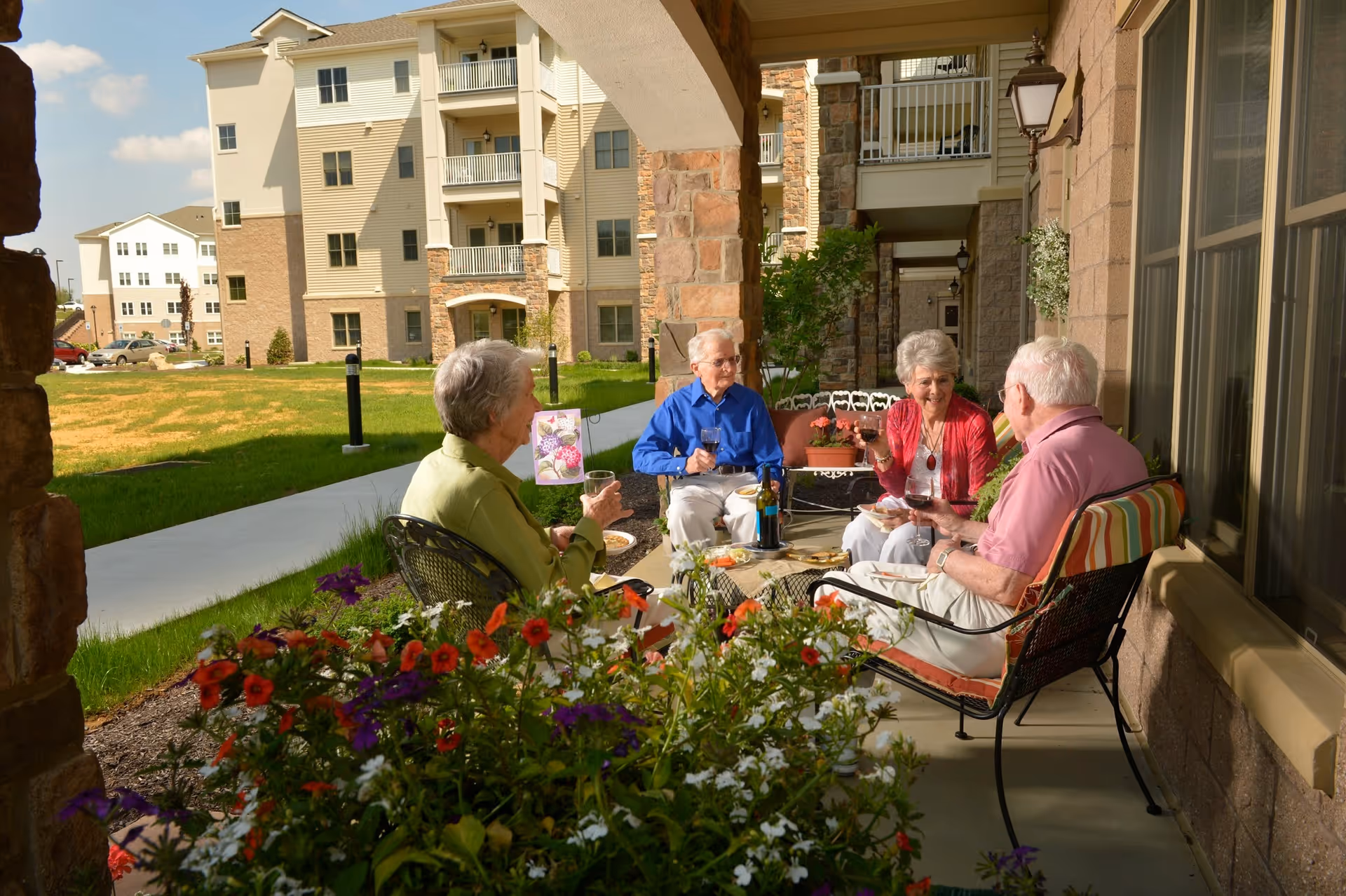 Four elderly people sitting and enjoying drinks together on a covered patio outside a senior living community building, surrounded by flowers and greenery.