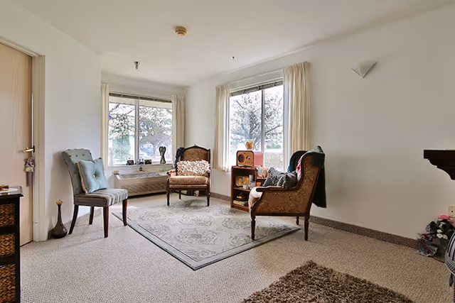 A cozy living room area with three vintage-style armchairs arranged around a patterned area rug. Large windows with cream-colored curtains let in natural light, and a small wooden shelf holds decorative items including a retro radio. The walls are painted white, and the carpet is light beige.