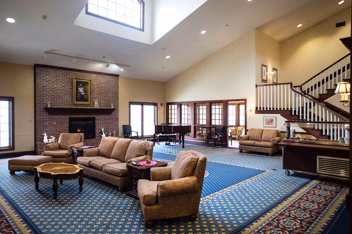 Spacious furnished common living area with sofas, armchairs, a fireplace, grand piano, and staircase under a skylight.