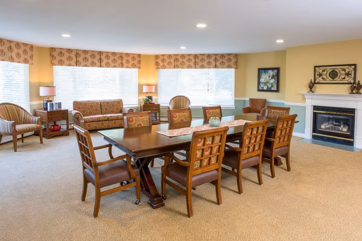 A spacious, well-lit common area with a large wooden dining table surrounded by eight cushioned chairs. The room features beige carpeting, yellow and green walls, and three large windows with patterned valances allowing natural light to fill the space. There are two armchairs and a patterned sofa near the windows, two side tables with lamps, a fireplace with decorative items on the mantel, and framed artwork on the walls.