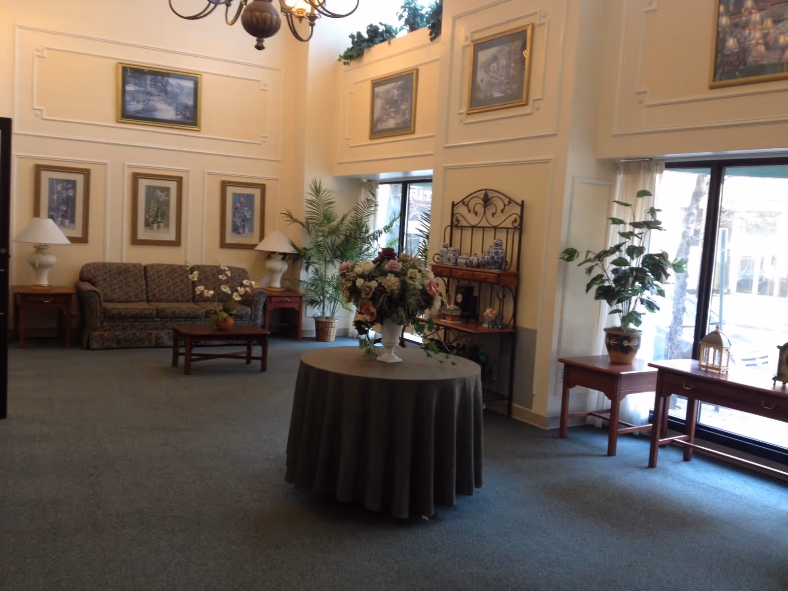 A spacious, well-lit living room area with a round table covered with a green tablecloth and a large floral arrangement in the center. There is a patterned sofa with two side tables and lamps, several framed paintings on the cream-colored walls, potted plants, and large windows letting in natural light. A decorative metal shelf with blue and white ceramics is also visible.