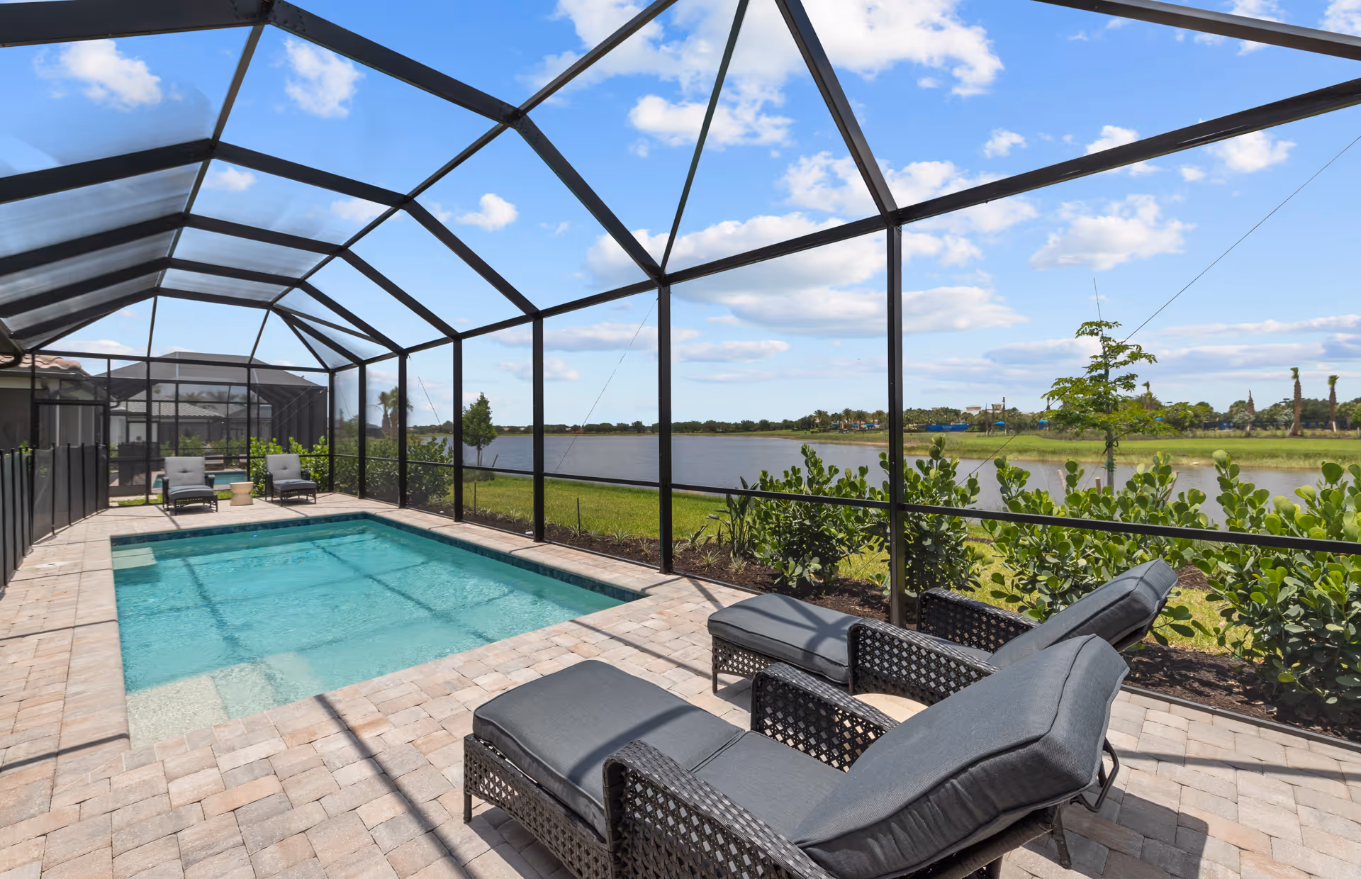 A screened-in outdoor pool area with two cushioned lounge chairs and two additional chairs near the pool. The pool is surrounded by a paved deck and overlooks a scenic view of a lake and greenery under a partly cloudy blue sky.