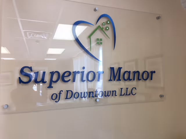 A clear acrylic sign mounted on a beige wall displaying the logo and name 'Superior Manor of Downtown LLC' with a blue and green house icon integrated into a heart shape above the text.