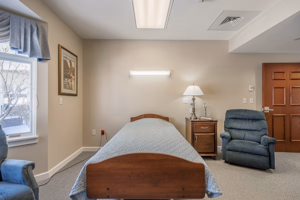 A simple bedroom in a senior living facility featuring a single wooden bed with a light blue quilt, a wooden nightstand with a lamp and some books, a blue upholstered armchair, a window with blue curtains, and a wooden door. The walls are beige and there is a framed picture hanging on the wall.