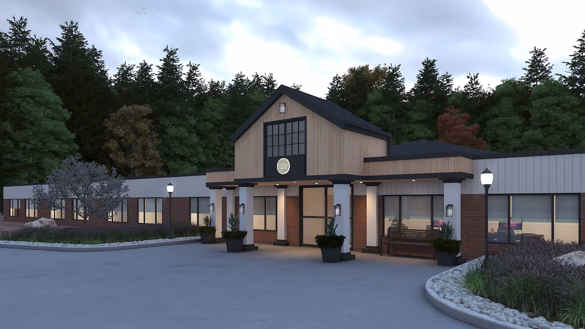 Exterior view of Lunabrook Village at Torrington showing the front entrance of a single-story building with a peaked roof section above the door, surrounded by trees and landscaped areas with plants and rocks under a cloudy sky.
