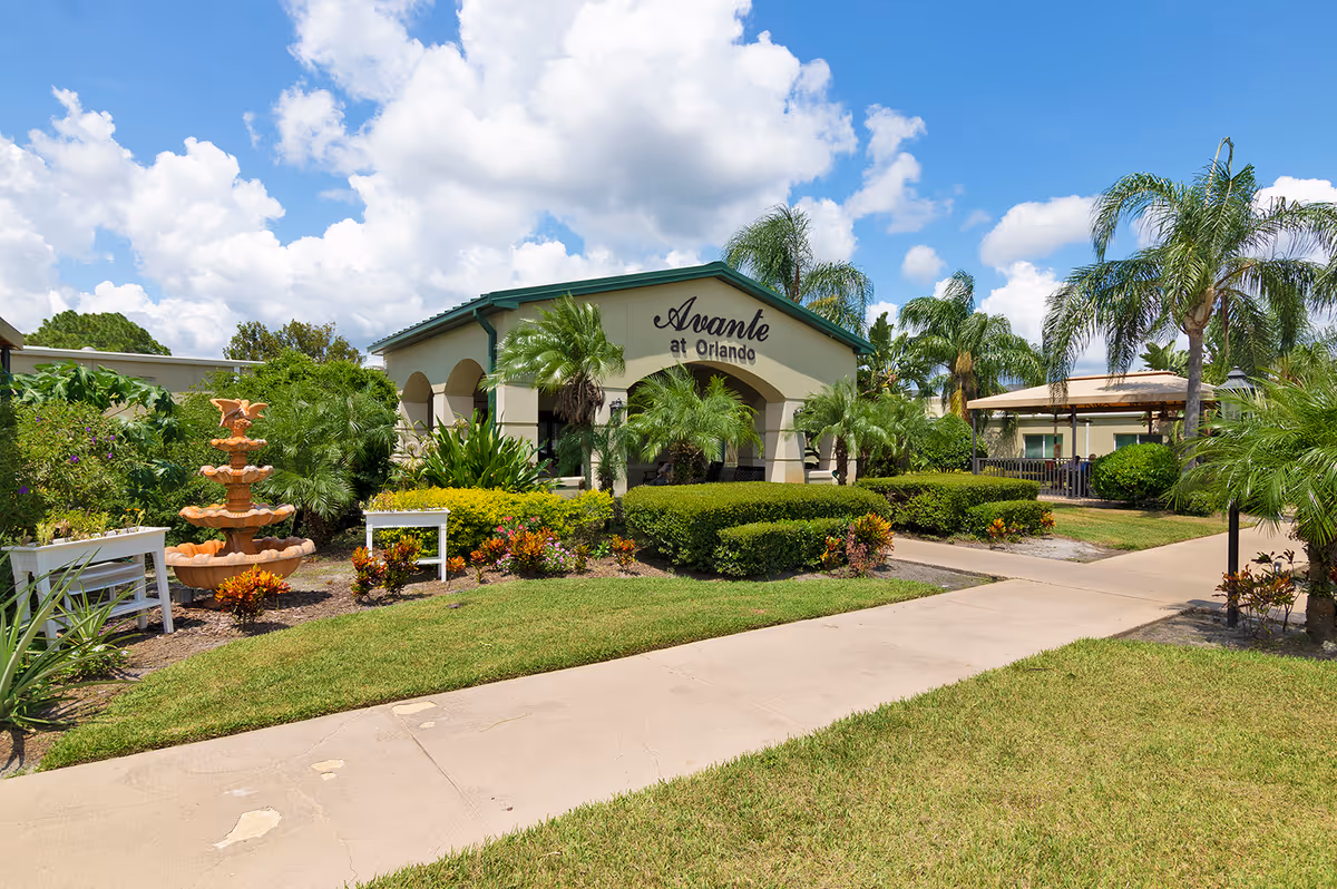 Exterior view of Avante at Orlando senior living facility with a clear blue sky and scattered clouds. The building has a green roof and beige walls with the name 'Avante at Orlando' displayed prominently. The foreground features a well-maintained garden with various plants, palm trees, a multi-tiered orange fountain, and a paved walkway leading to the entrance.