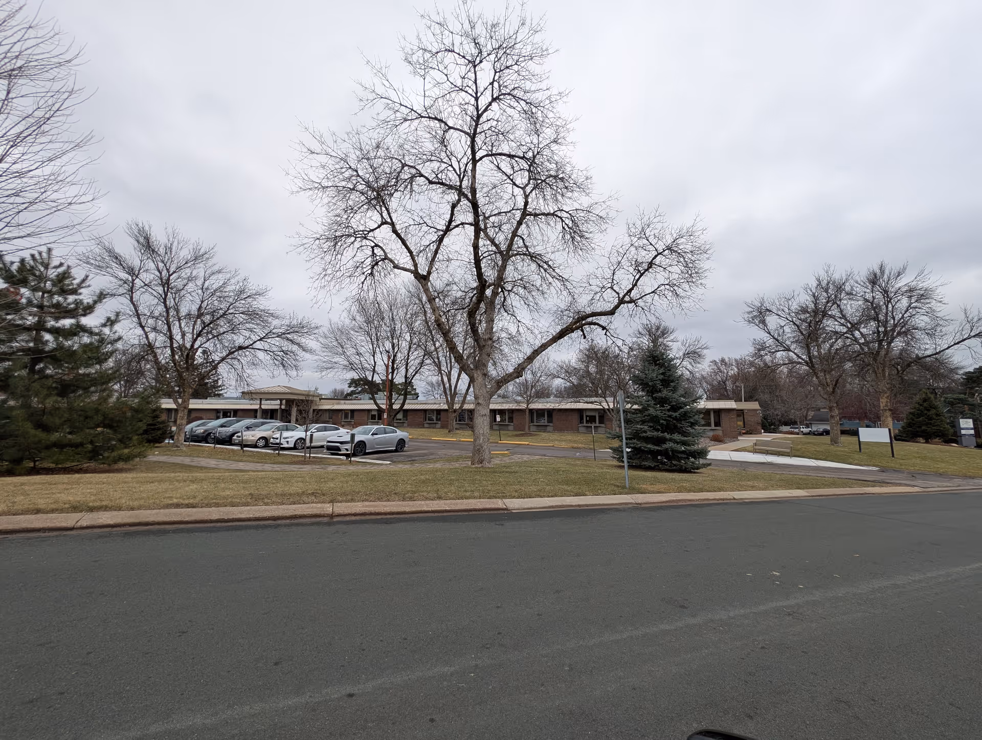 Single-story senior living building with parked cars, leafless trees, and a lawn under an overcast sky.