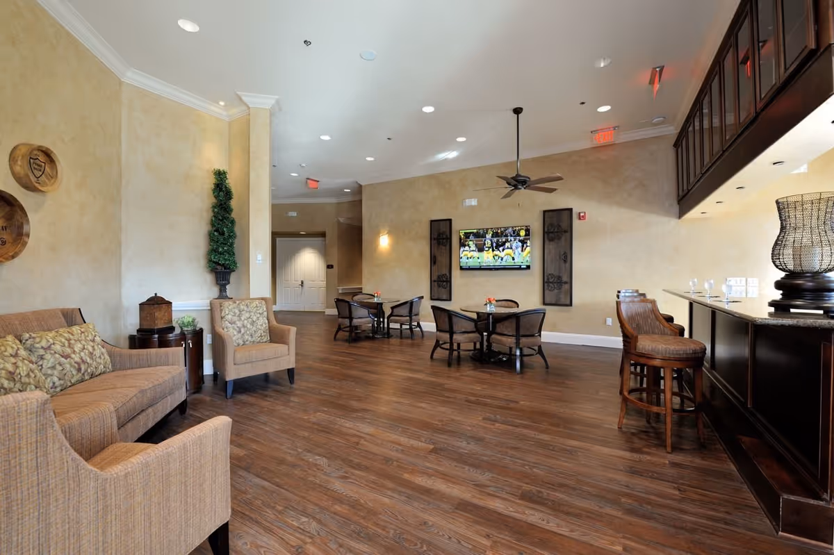 A spacious living room area with wooden flooring, beige walls, and recessed lighting. The room features a sofa and armchairs with patterned cushions on the left, two small round tables with chairs in the center, and a dark wood bar with high stools on the right. A flat-screen TV is mounted on the far wall, displaying a football game. Decorative wall art and a tall potted plant add to the cozy ambiance.